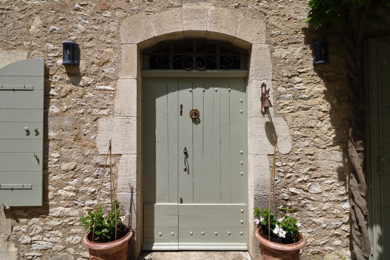 An elegant light gray door with decorative ironwork framed by a stone archway, flanked by two potted plants and shaded by wooden shutters on a sunny day.